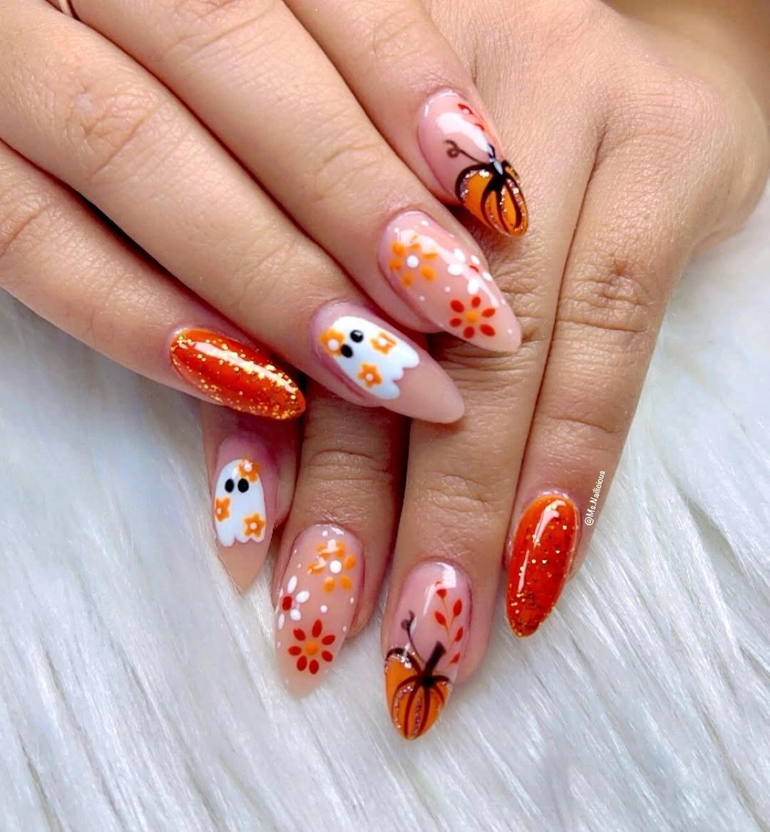 Soft nude almond nails with pumpkin, white flowers, orange details, and glitter accent