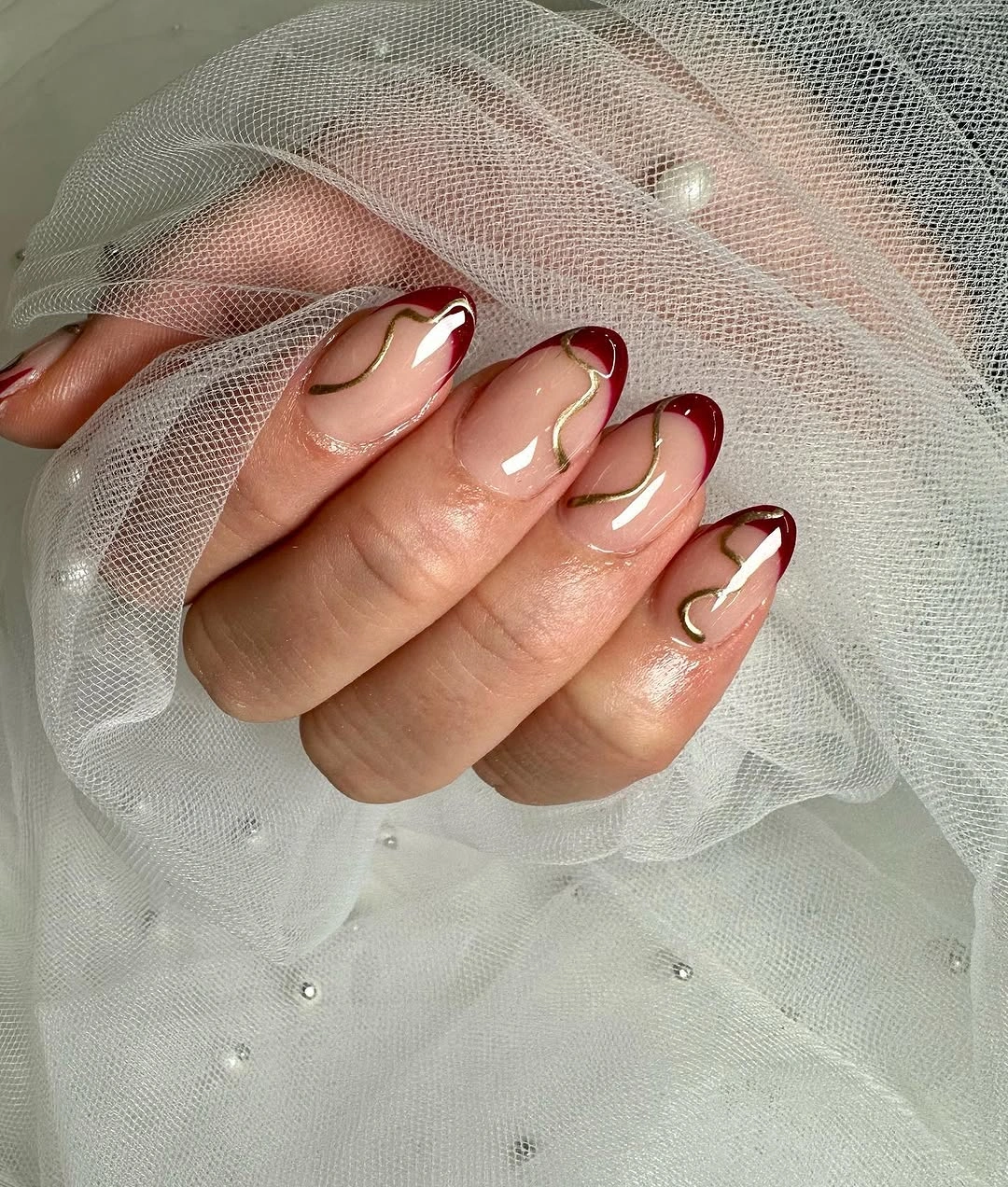 Sheer nude almond nails with slim burgundy French tips and fine gold curves