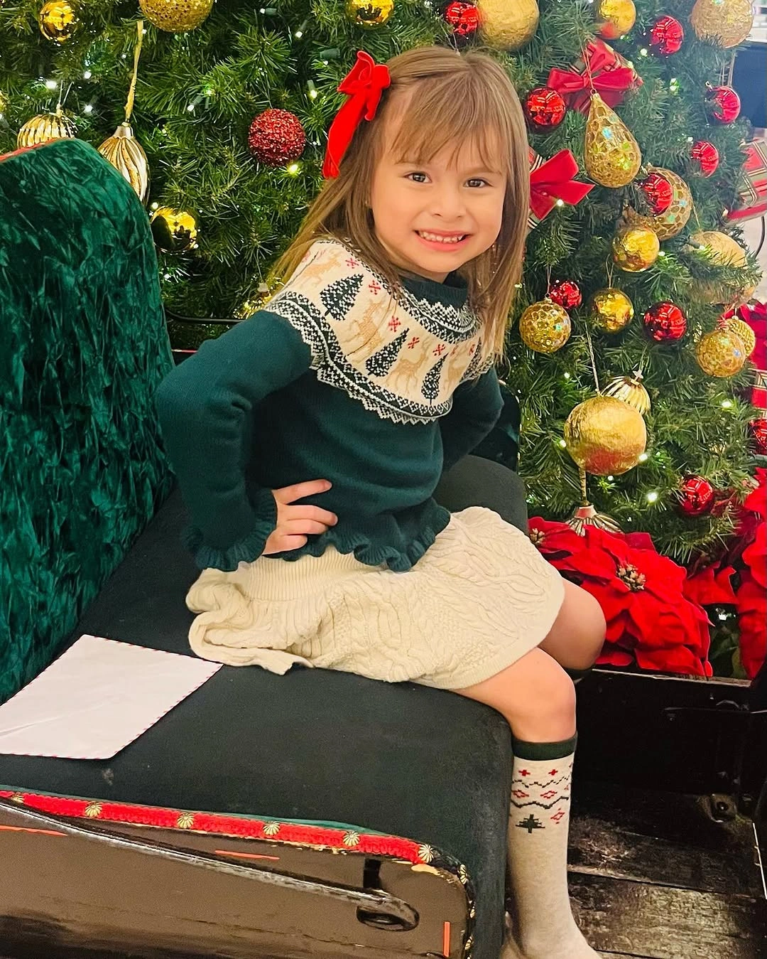 Child in green sweater and white skirt beside Christmas tree