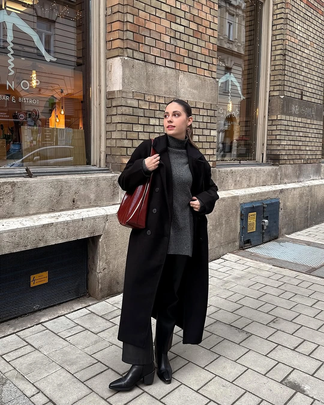 Long black coat with charcoal knit, wide trousers, black boots, and burgundy bag