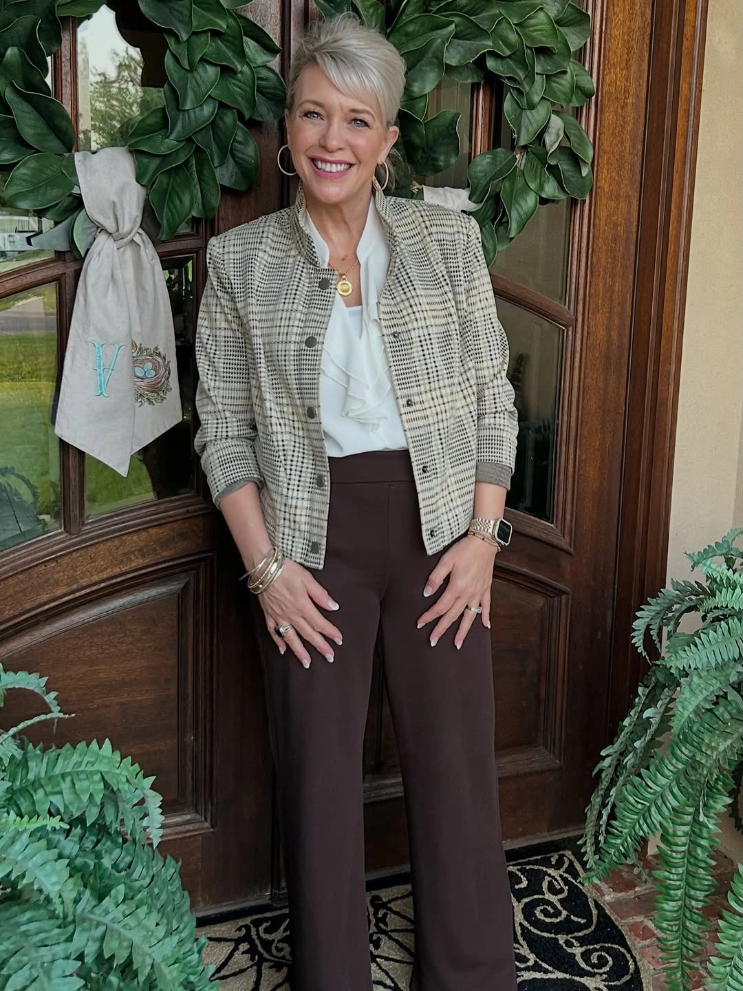 Short checked jacket over a pale ruffled blouse with dark brown trousers.
