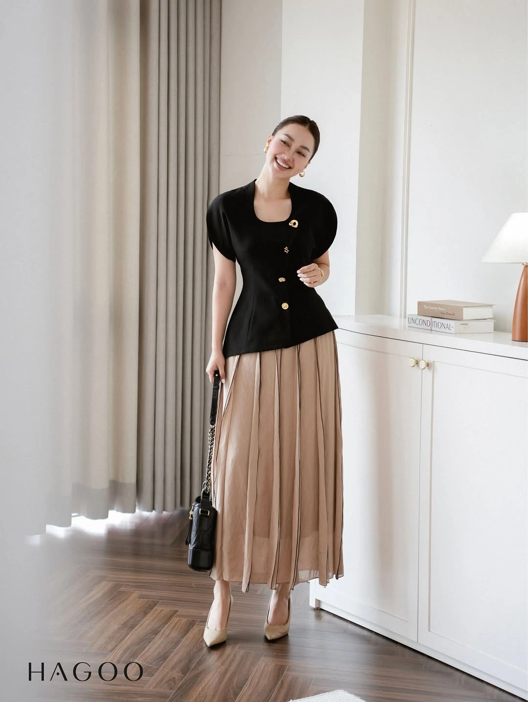 Black waist-shaped top with gold-colored buttons, beige midi-length pleats, and pale pointed shoes.