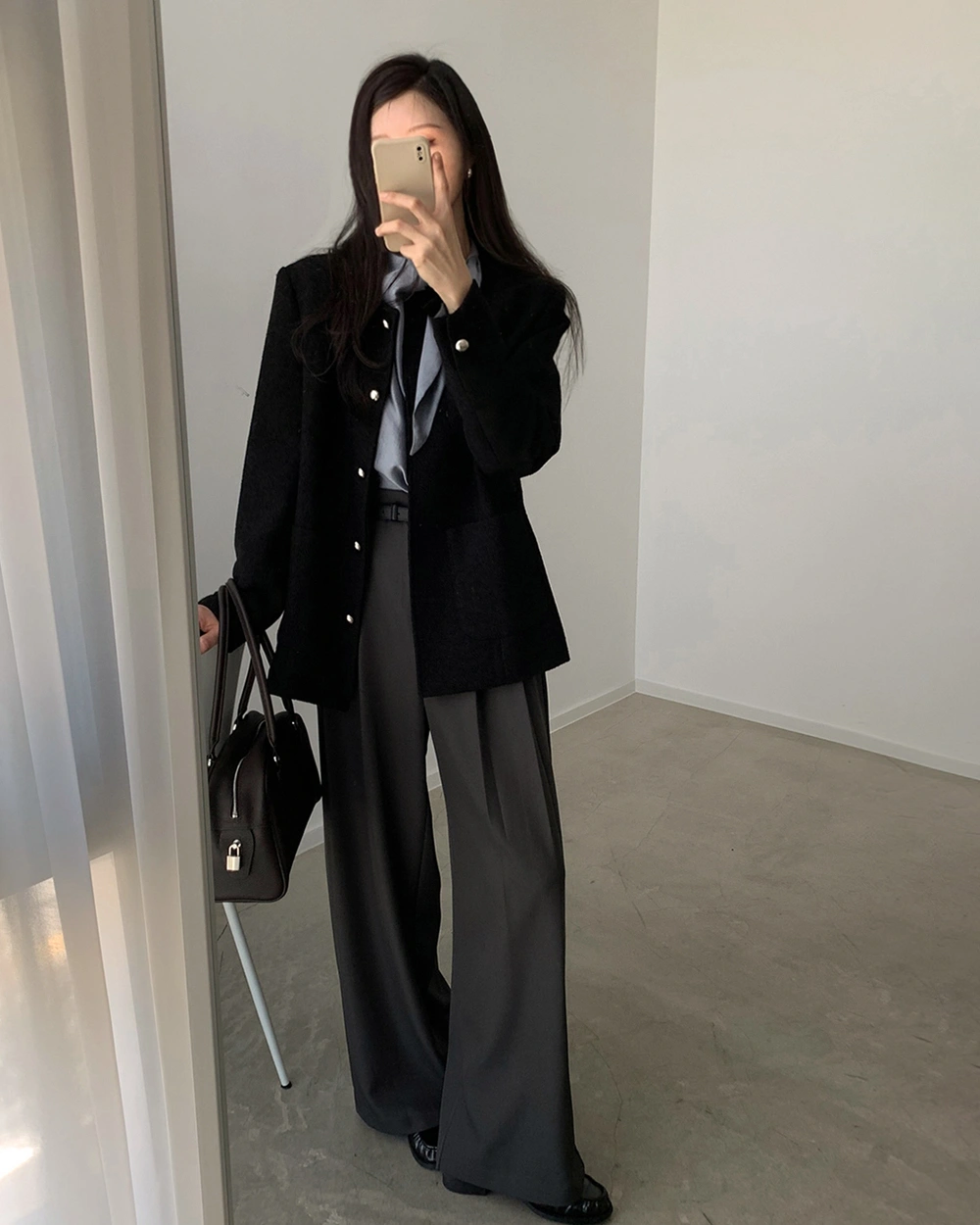 Long black jacket, gray blouse, wide charcoal trousers, and structured black handbag.
