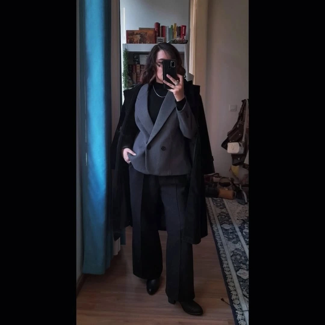 Gray double-breasted blazer beneath a draped black coat, with dark wide-leg trousers.