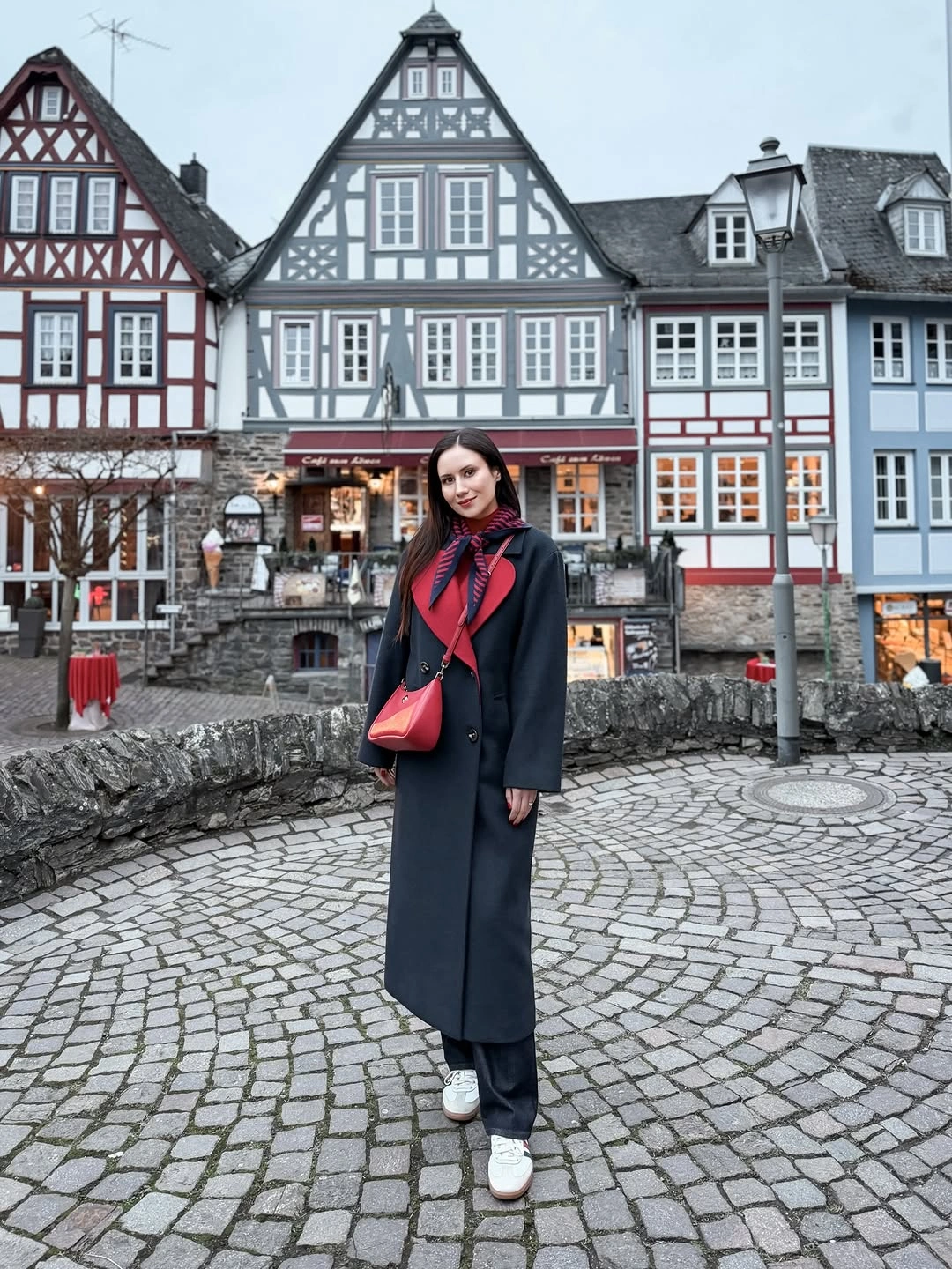 Dark long coat with red sweater, scarf, bag, trousers, and white sneakers