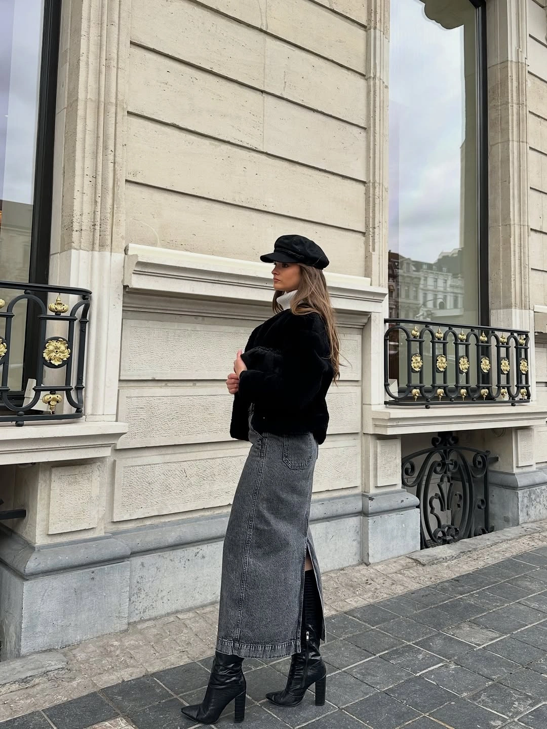 Black textured cropped jacket with a gray denim midi skirt, cap, and heeled knee boots