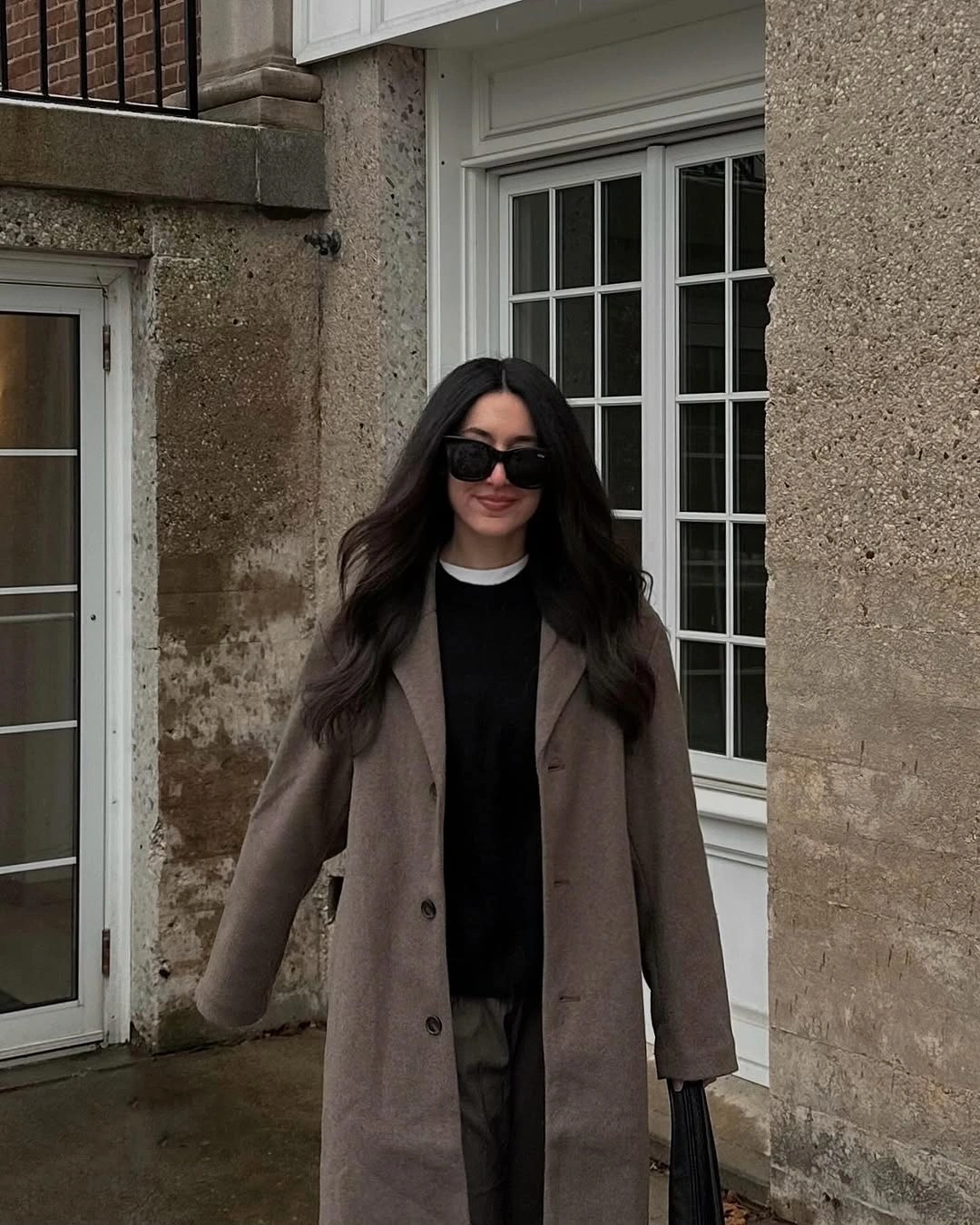 Taupe-brown long coat, black top, dark pants, sunglasses, and bag
