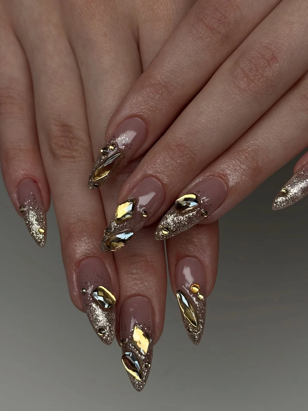 Long pointed nude nails with gold-colored reflective pieces and glitter