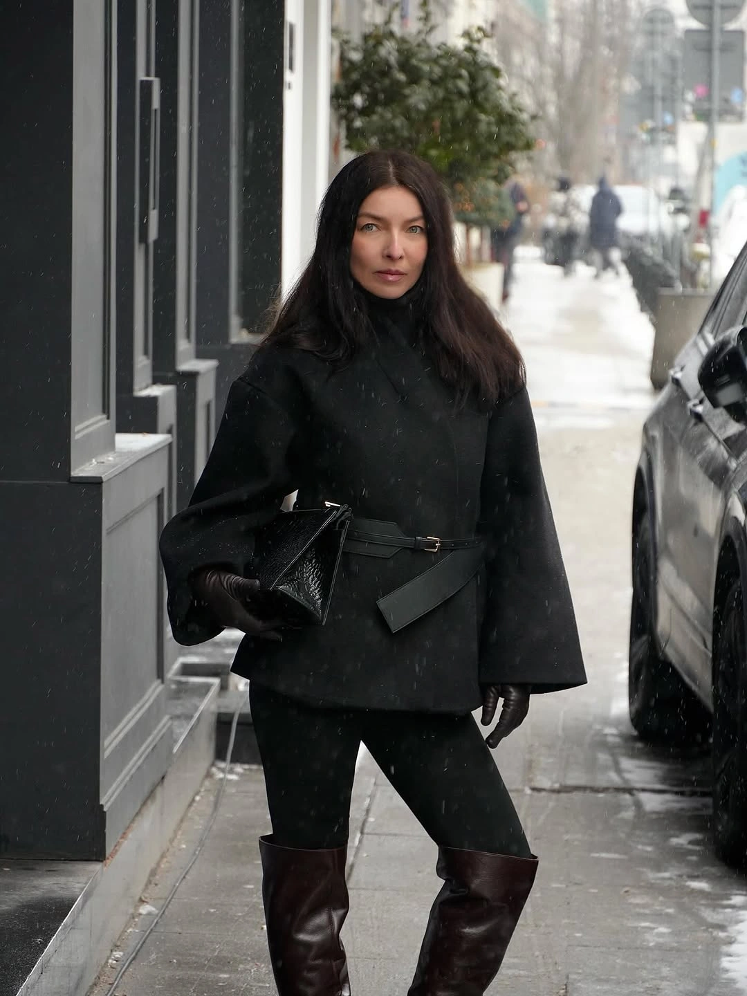 Black cape-style jacket black pants knee-high brown boots and angular black clutch