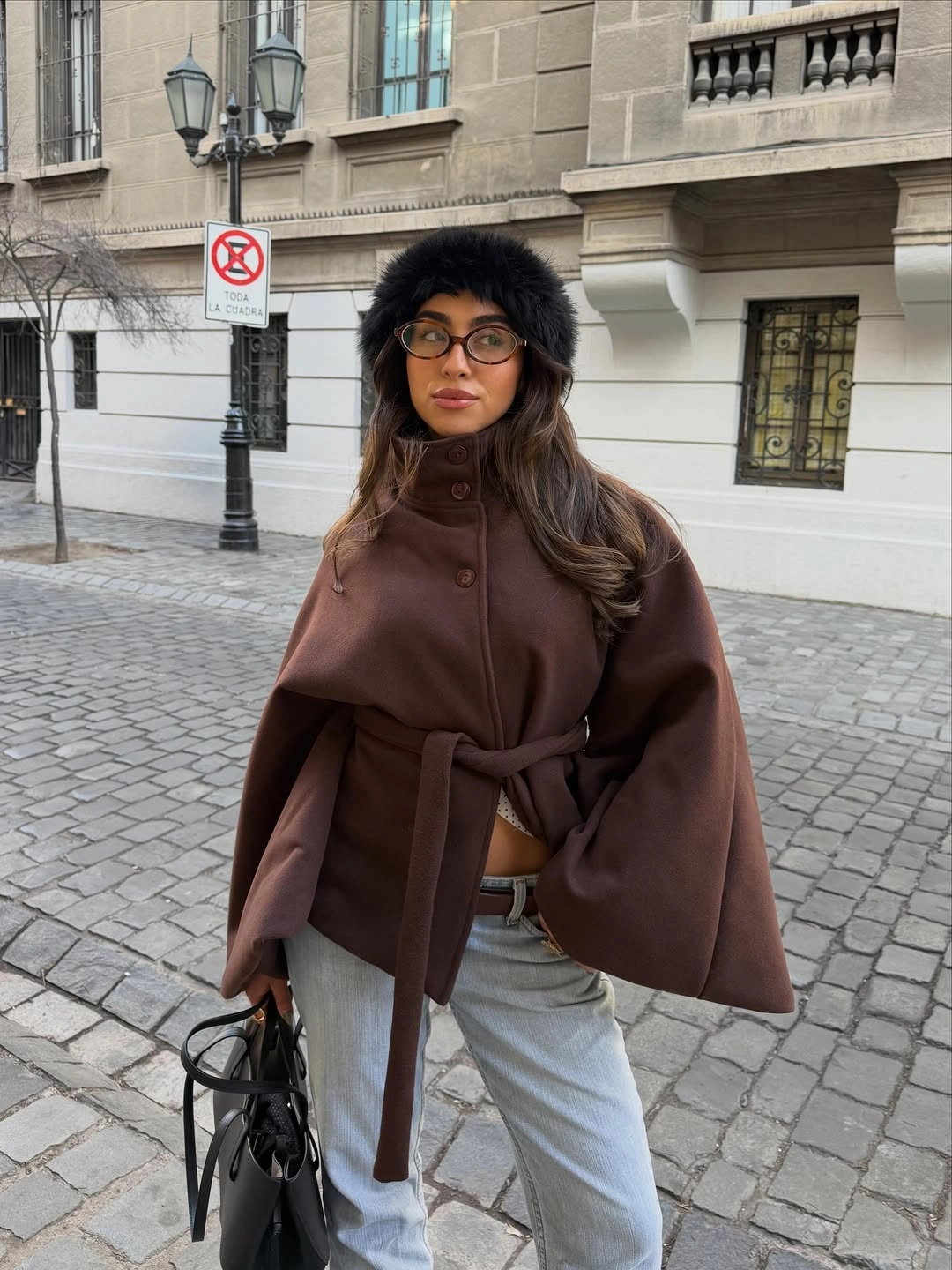 Belted brown cape-style jacket with light blue jeans black furry hat and handbag