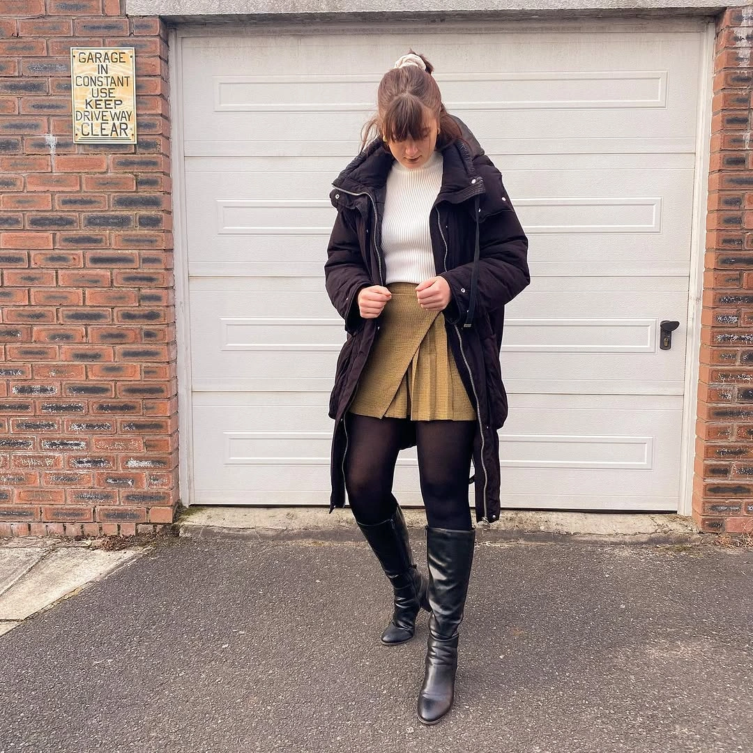 Black mid-length puffer with cream sweater, mustard skirt and tall boots