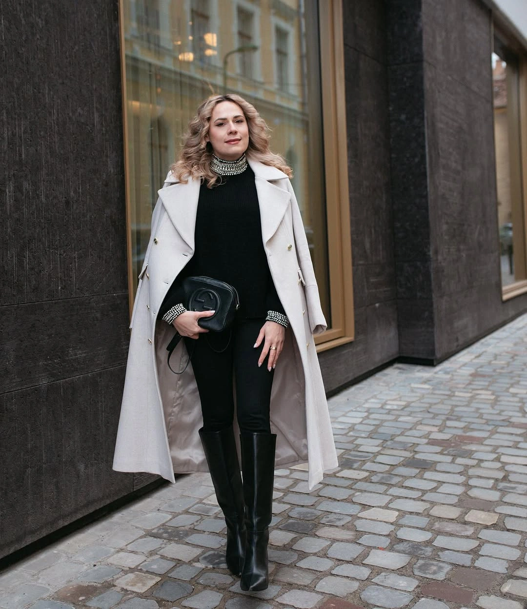 Pale double-breasted coat over black knit, trousers, and knee-high boots