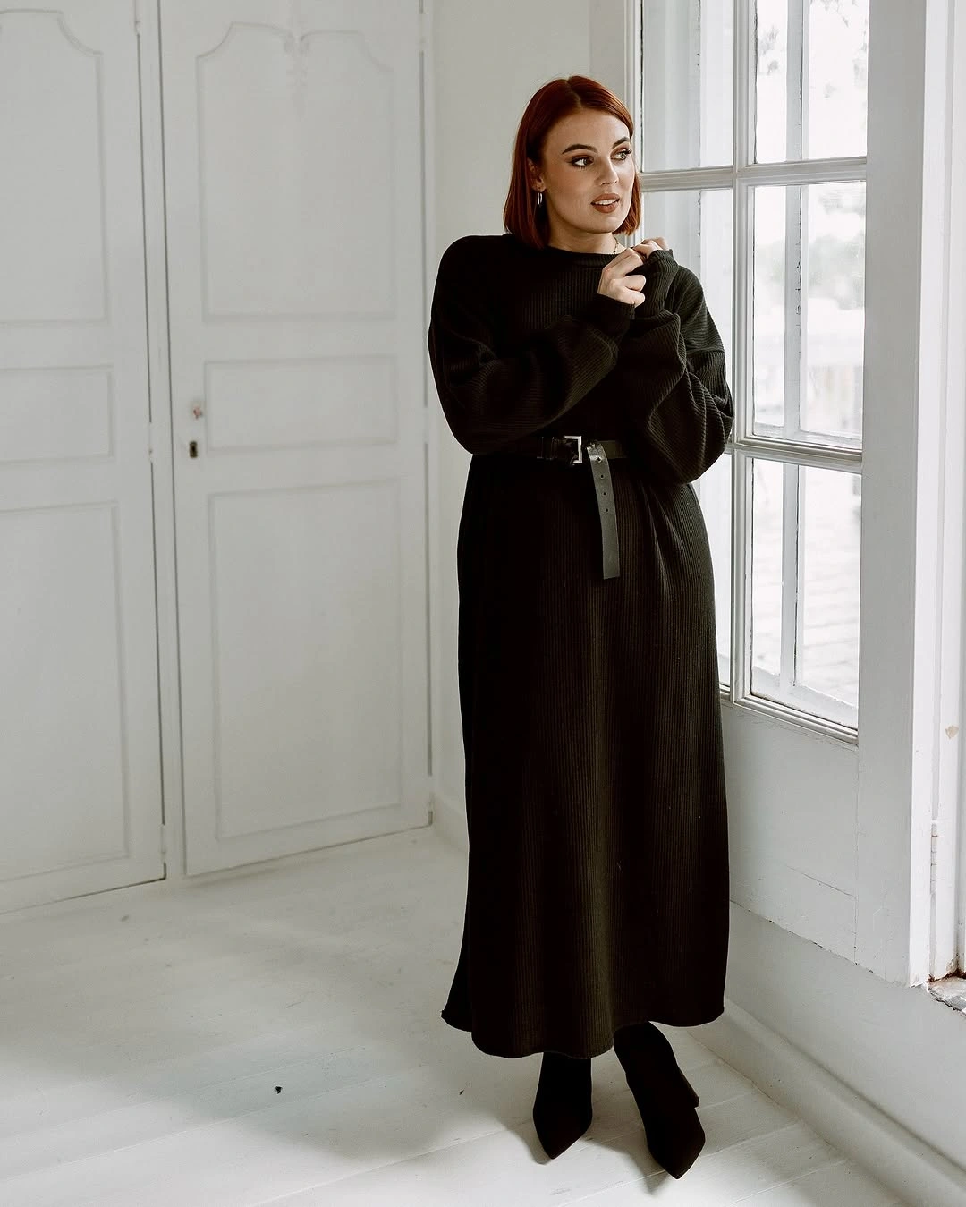 Long dark belted dress with black pointed ankle boots indoors