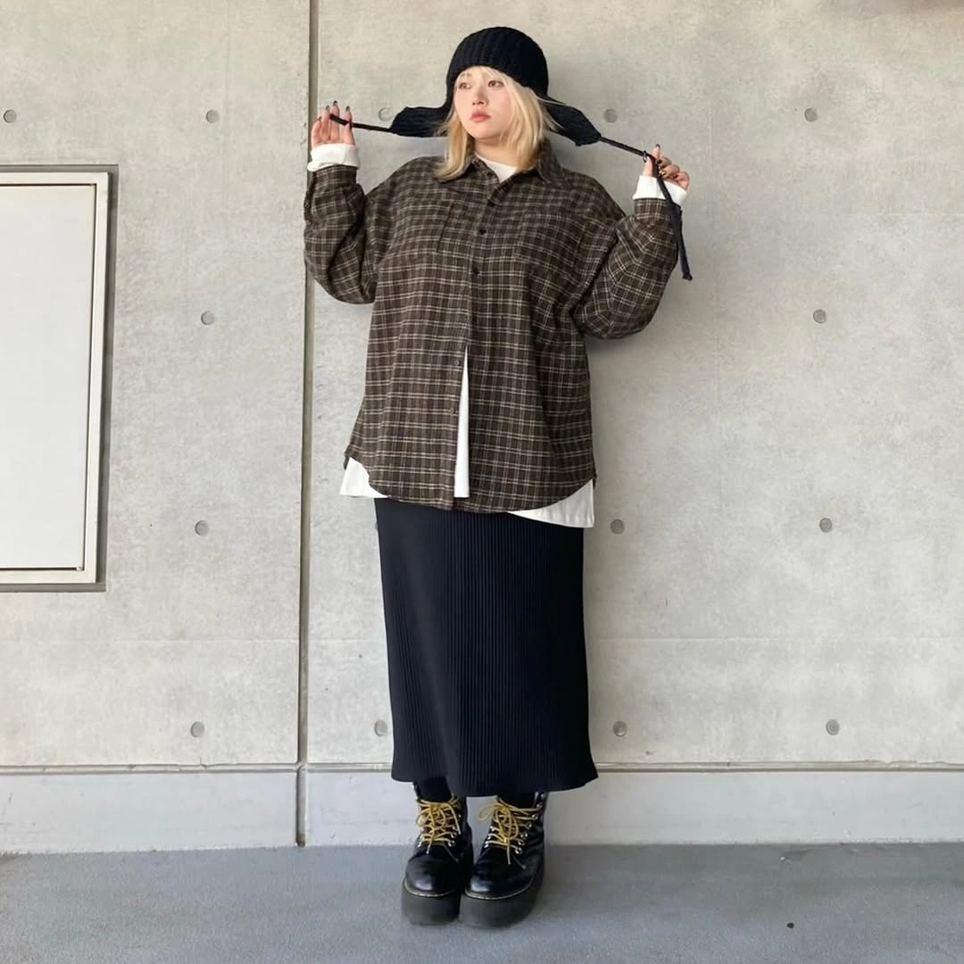 Person in oversized plaid shirt, long dark skirt, hat, and boots