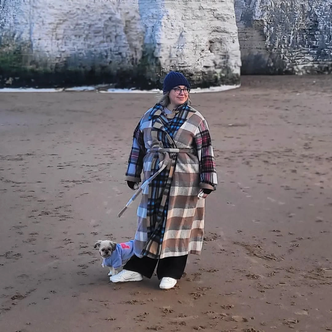 Person in long plaid coat, blue scarf, beanie, and white sneakers