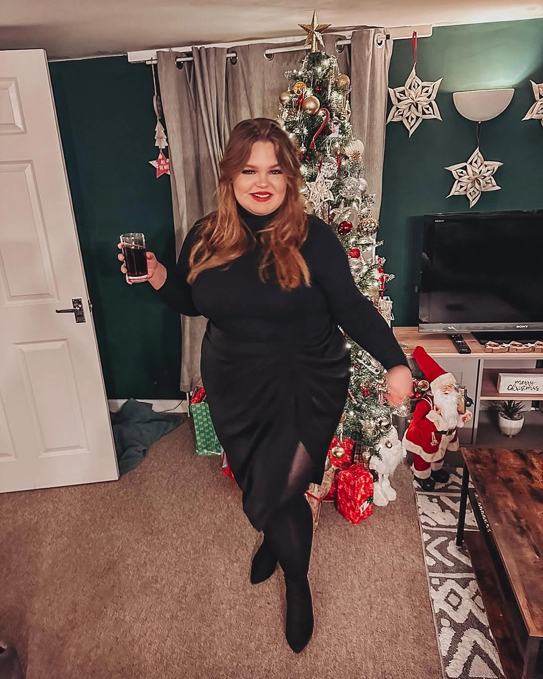 Woman in black dress, black boots, red lipstick, and decorated tree