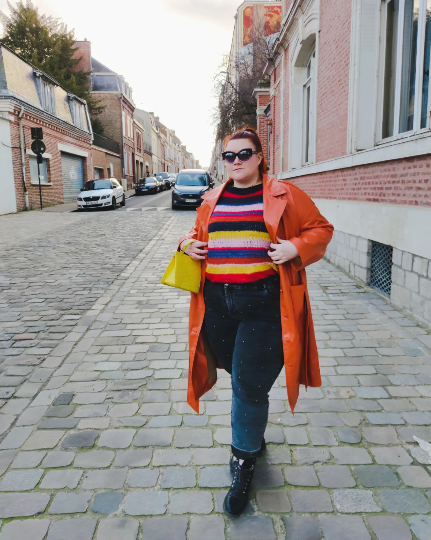 Woman in orange coat, striped sweater, dark jeans, and yellow bag