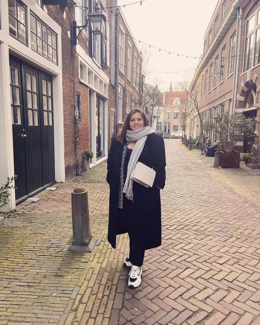 Woman in long black coat, pale scarf, light bag, and sneakers
