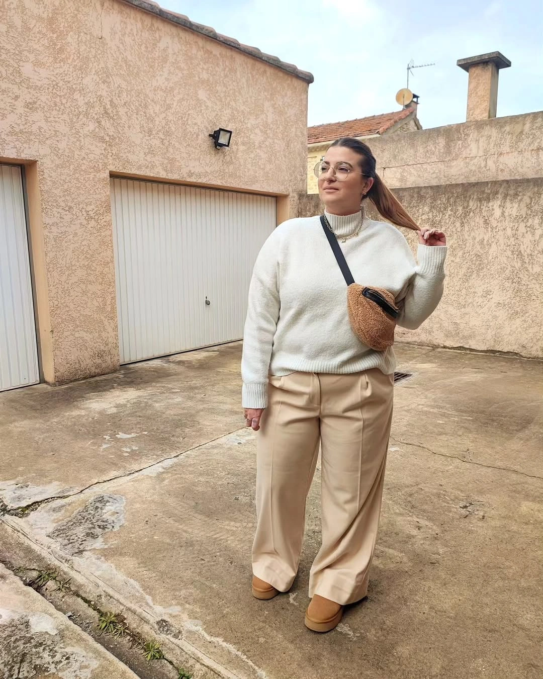Woman in cream sweater, beige trousers, tan shoes, and sling bag