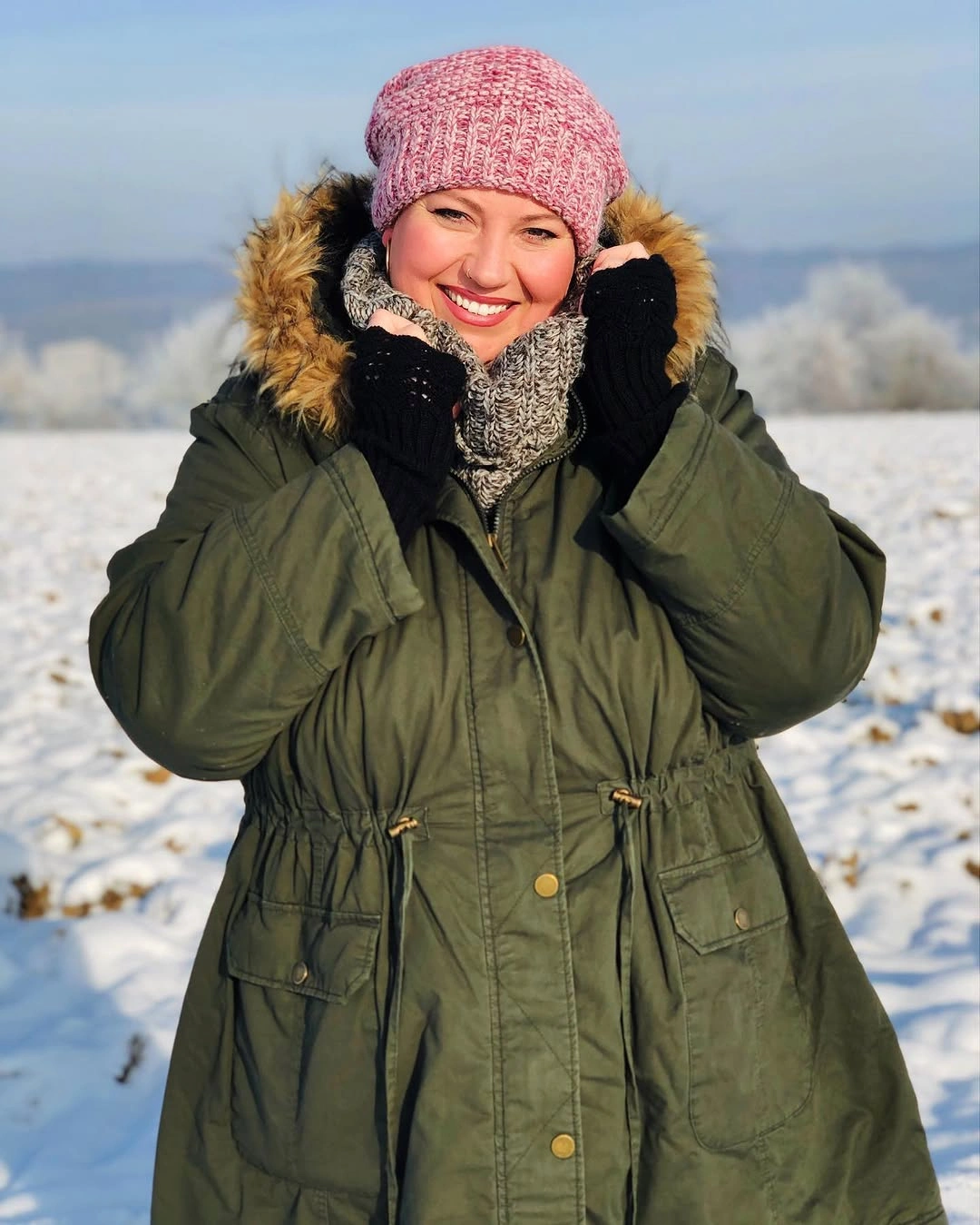 Woman in olive hooded parka, gray scarf, and pink beanie