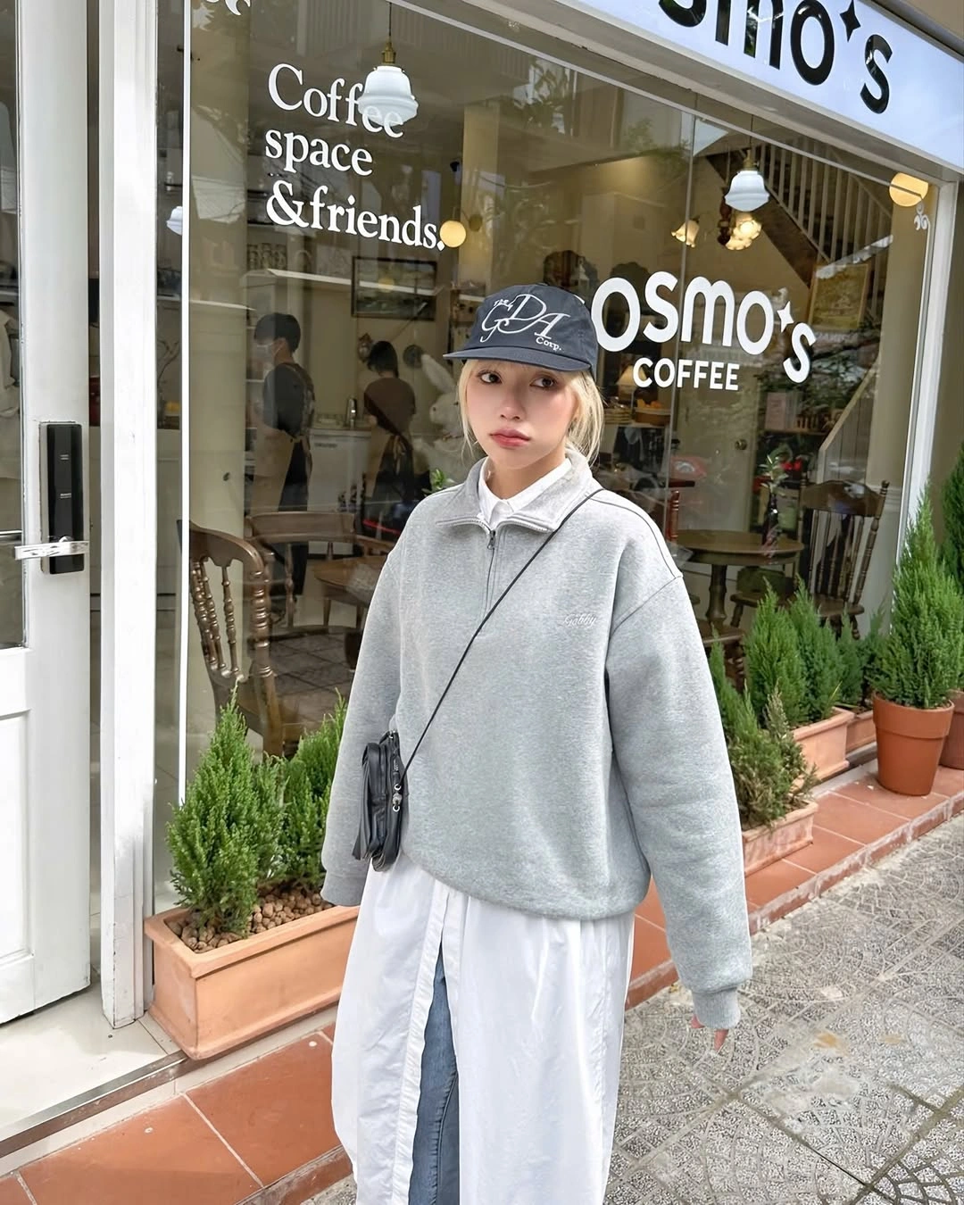 Gray quarter-zip pullover, long white shirt, blue-gray trousers, cap, and crossbody