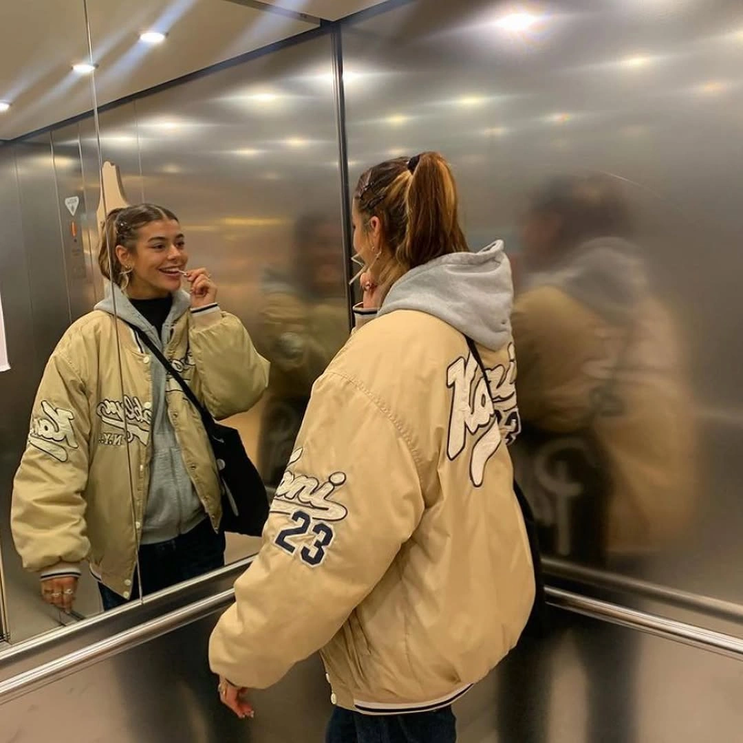 Beige padded varsity jacket over gray hoodie with black shoulder bag