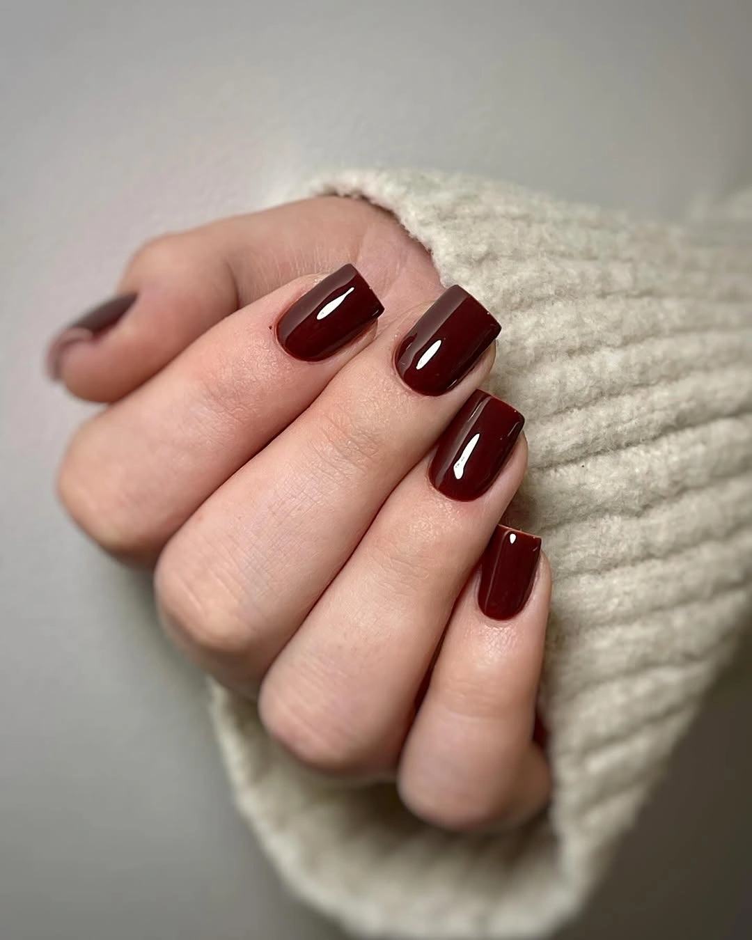 Short glossy burgundy square nails beside cream knit sleeve