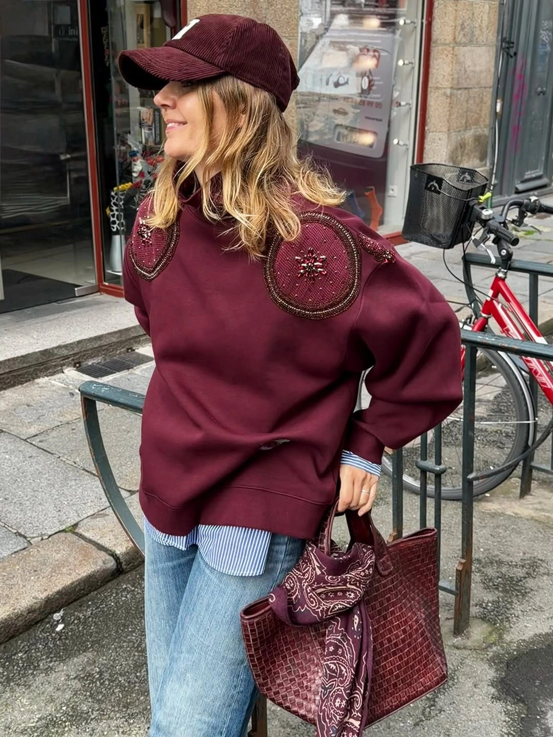 Burgundy cap and embellished sweatshirt with striped shirt, blue jeans and handbag
