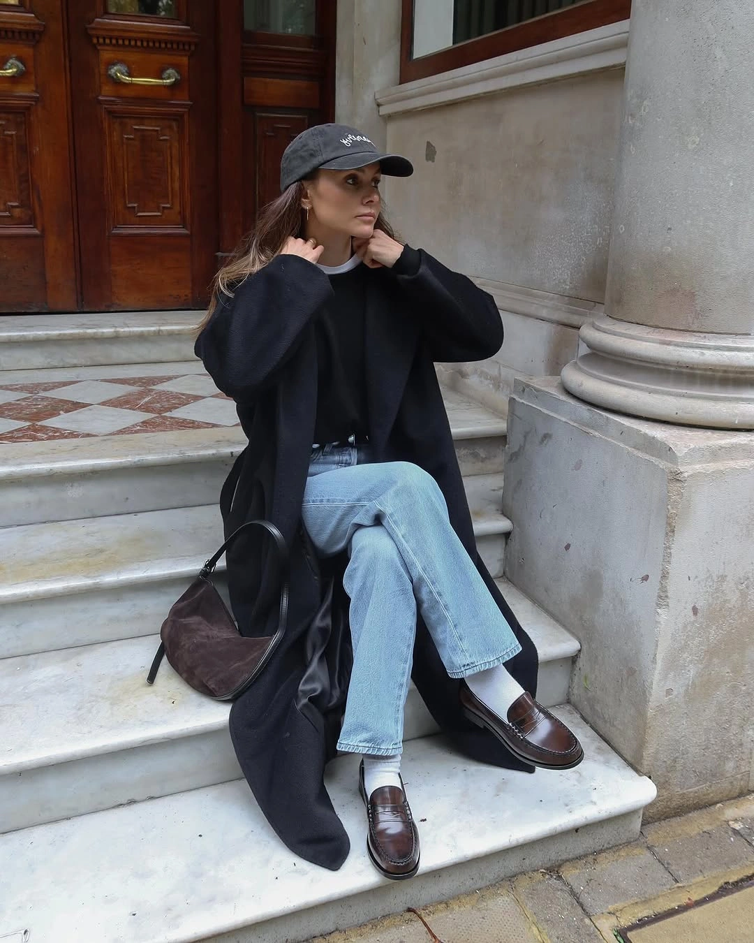 Woman in long black coat, light jeans, cap, and loafers