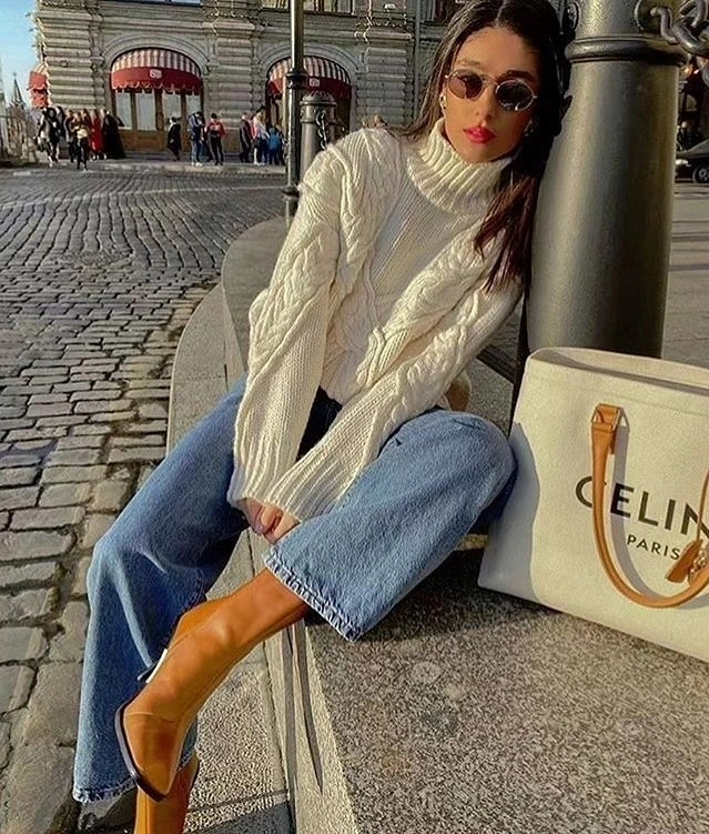 Woman in cream cable-knit sweater, blue jeans, and tan boots