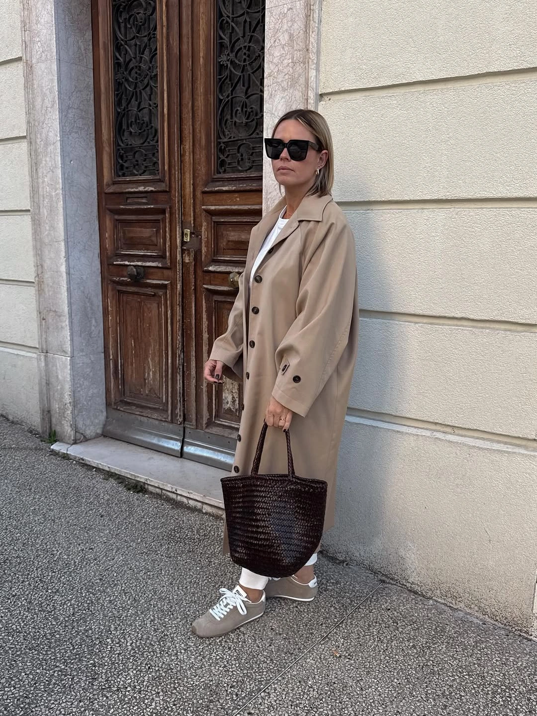 Light taupe trench coat with neutral sneakers and dark woven handbag