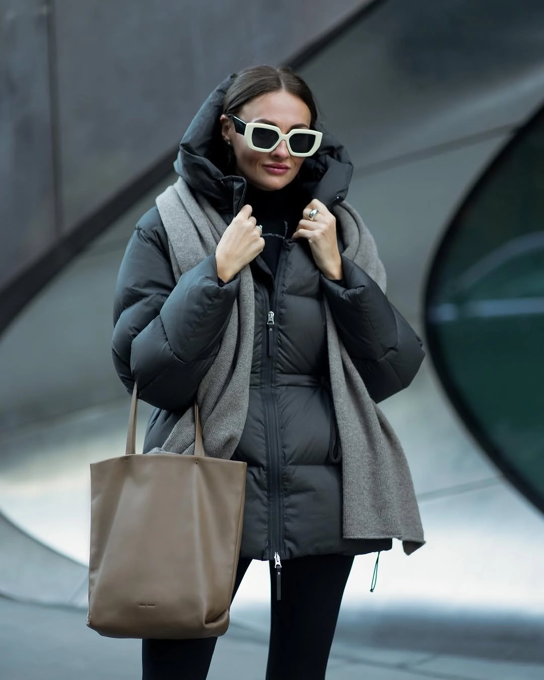 Woman in black hooded puffer jacket, gray scarf, white sunglasses and taupe tote