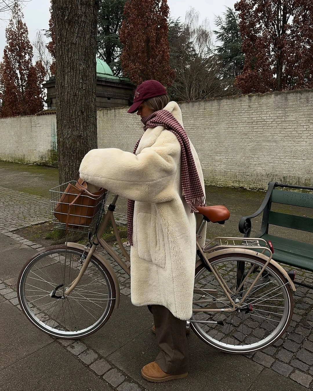 Woman in long cream plush coat, burgundy cap and checked scarf beside bicycle