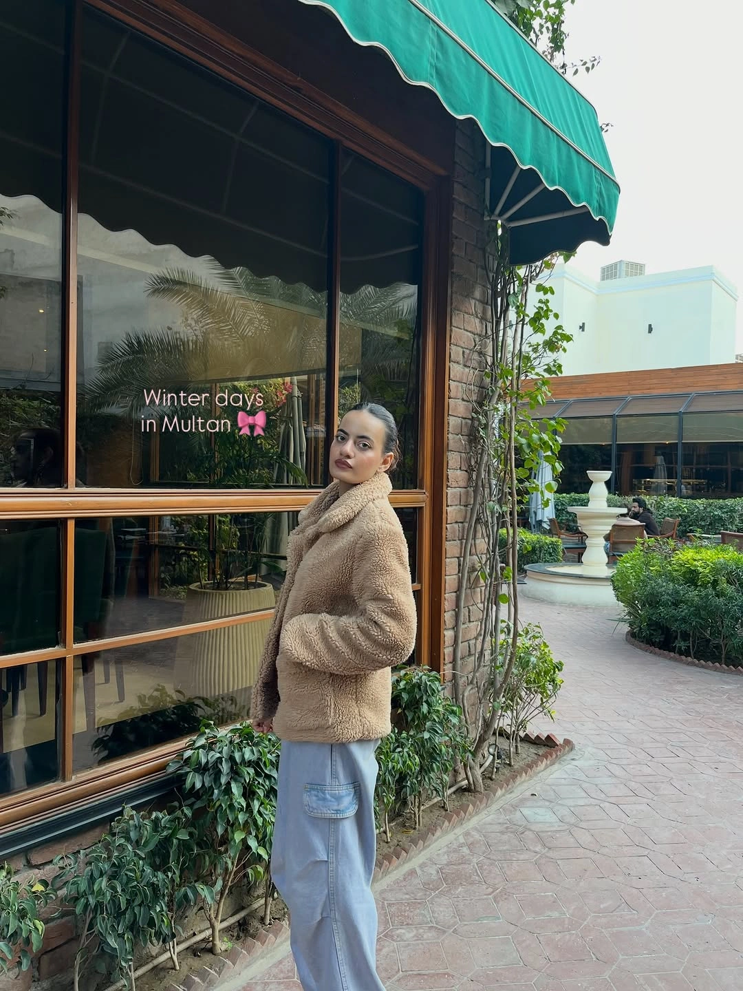 Woman in tan textured jacket and loose pale blue cargo trousers outside cafe