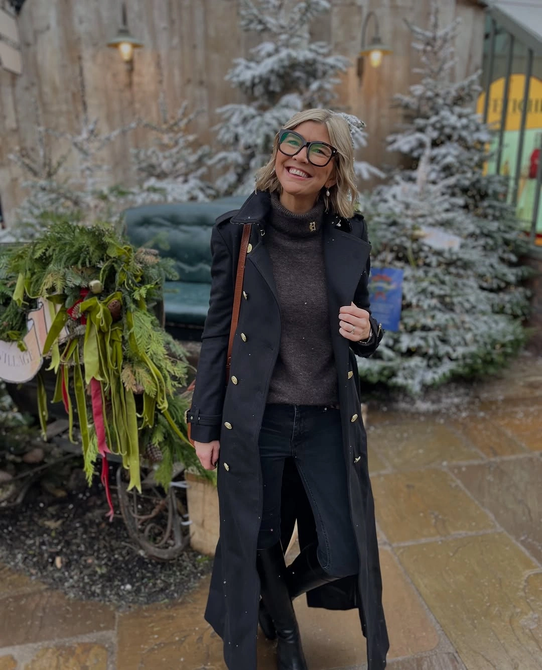 Smiling woman in long dark coat, gray high-neck sweater and green glasses