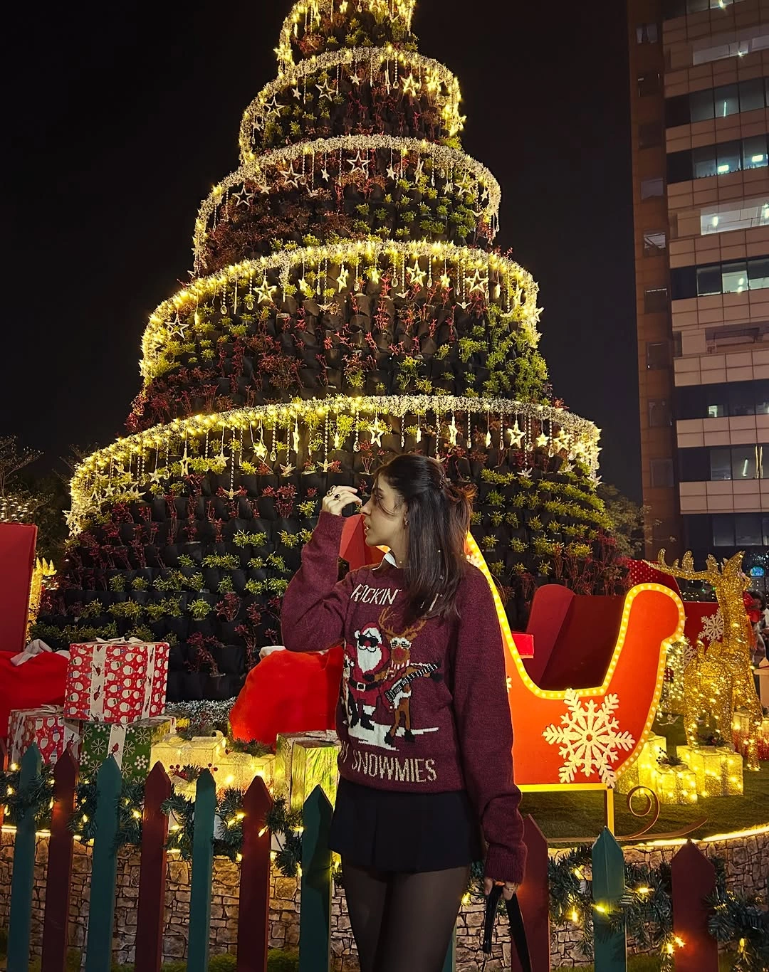 Woman in burgundy graphic holiday sweater, short dark bottoms and tights beside illuminated tree