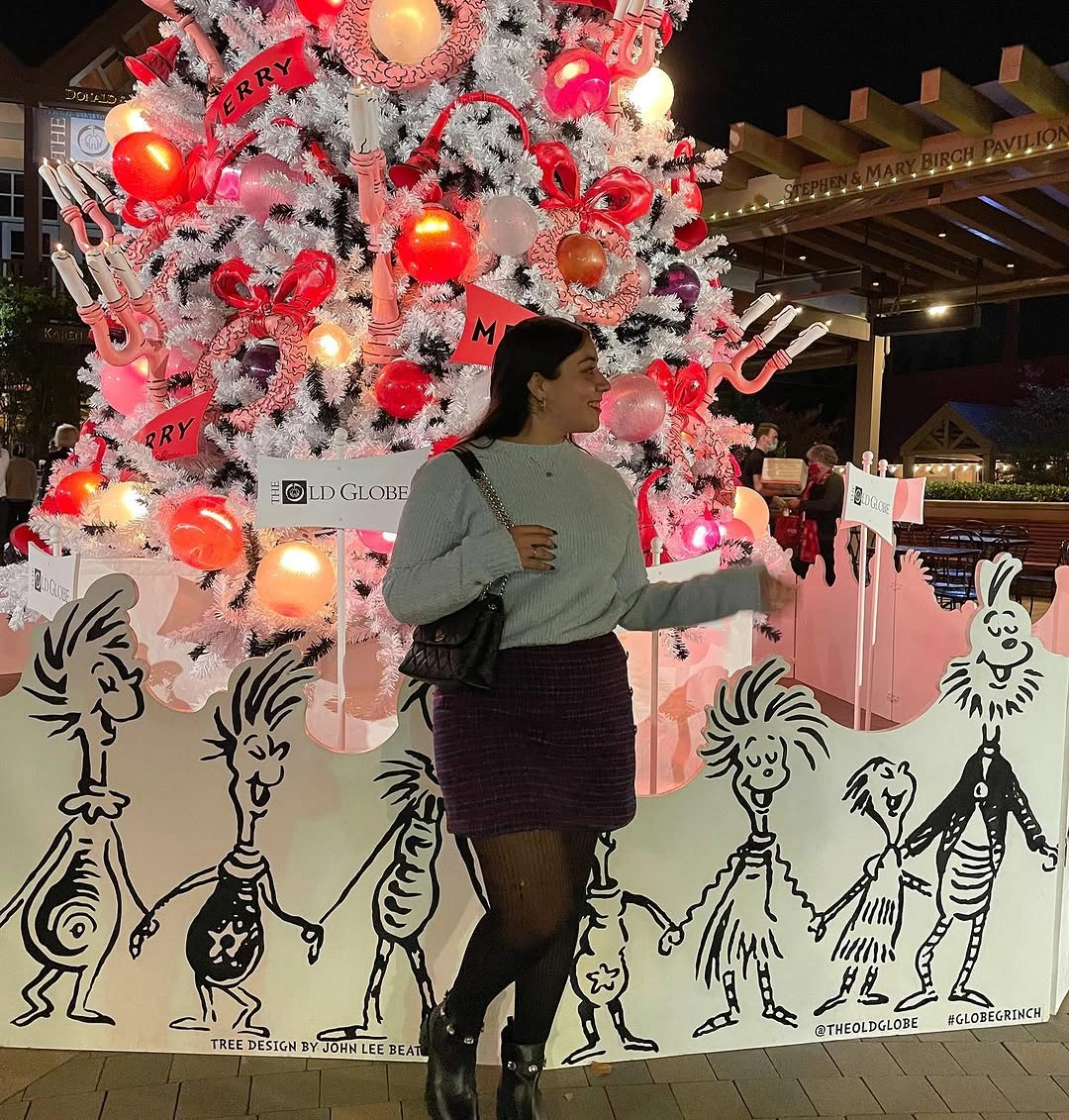 Woman in pale sweater, dark plaid mini skirt, tights and boots beside Christmas tree