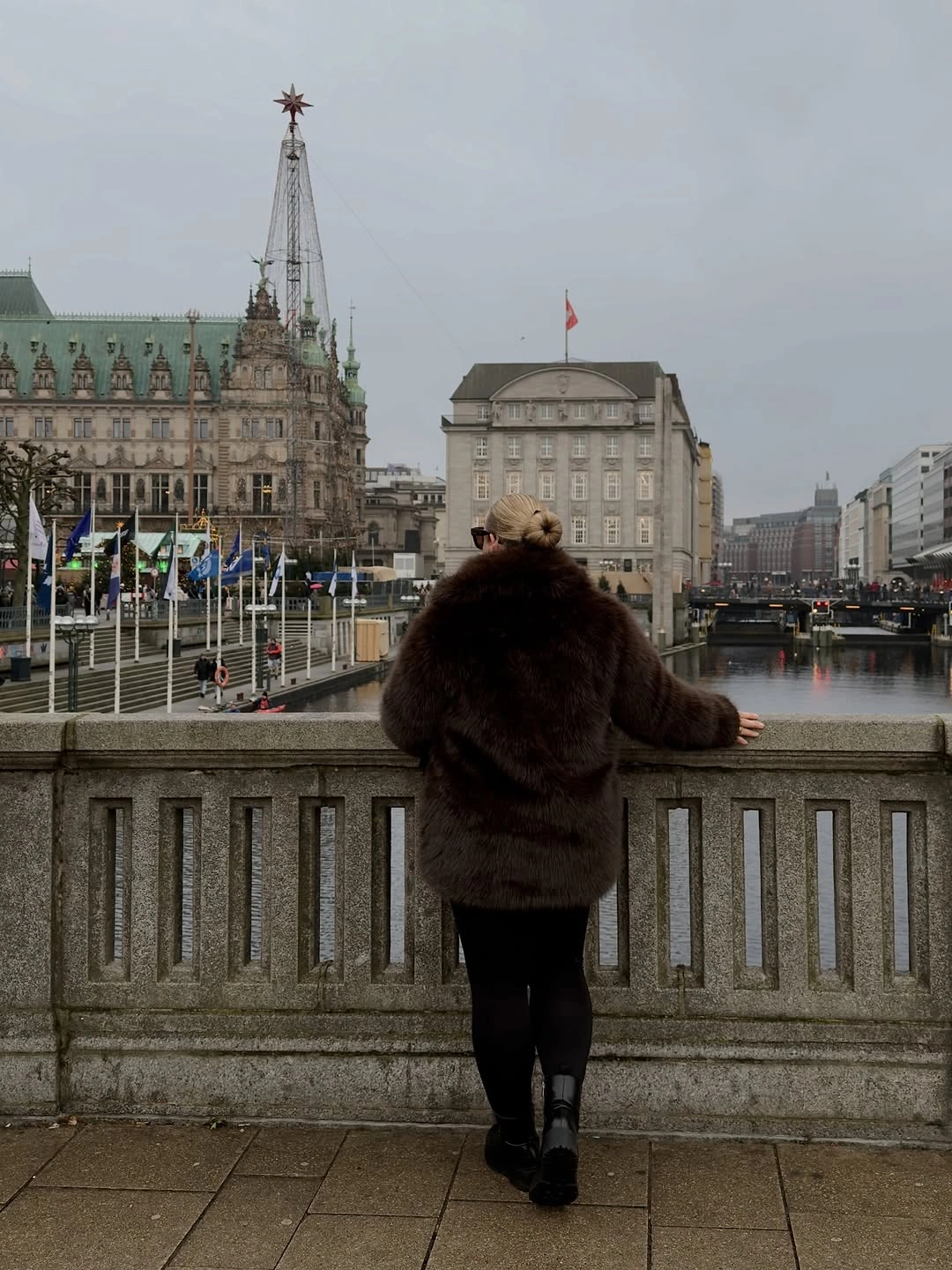 Woman viewed from behind in plush brown jacket, black pants and boots at bridge