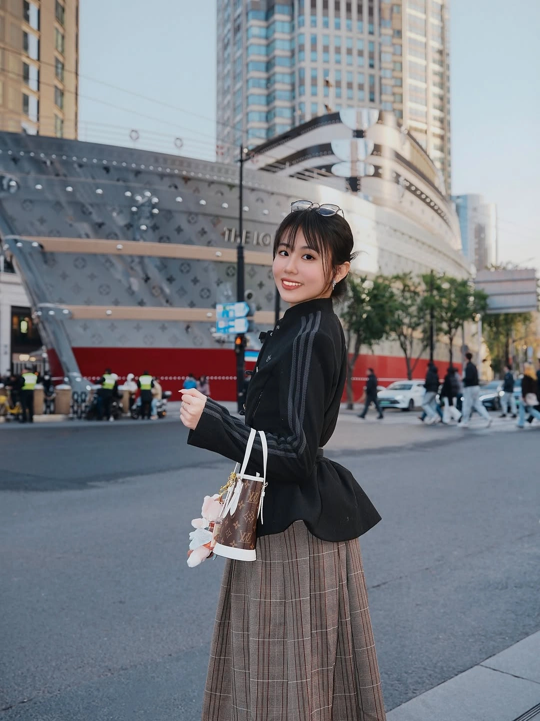 Woman in fitted black jacket and long brown plaid skirt carrying patterned handbag