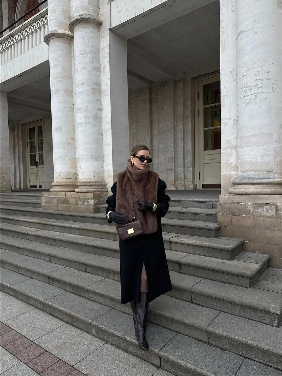 Long black coat with broad brown plush layer, dark boots, and handbag