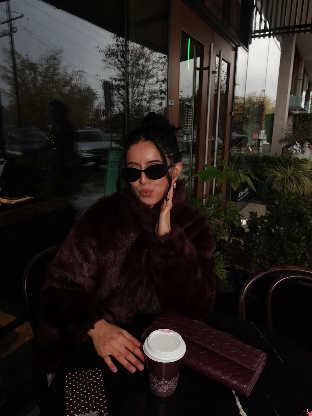 Dark plush jacket, burgundy quilted bag, black sunglasses, and takeaway cup