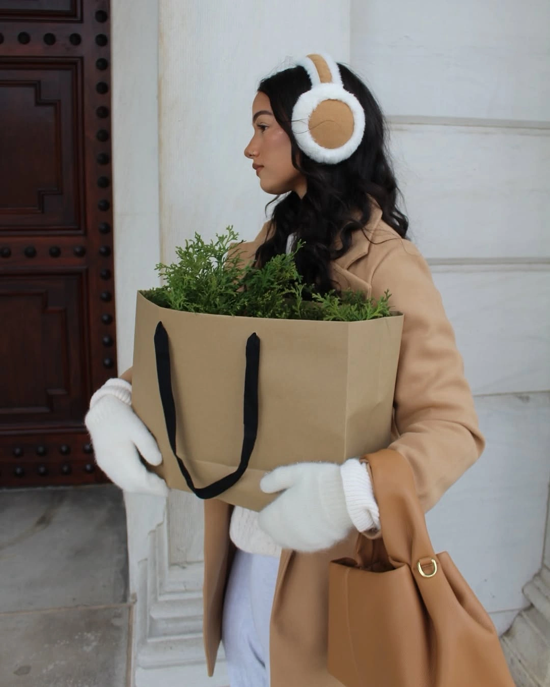 Camel coat with white earmuffs, white mittens, pale trousers, and tan bag