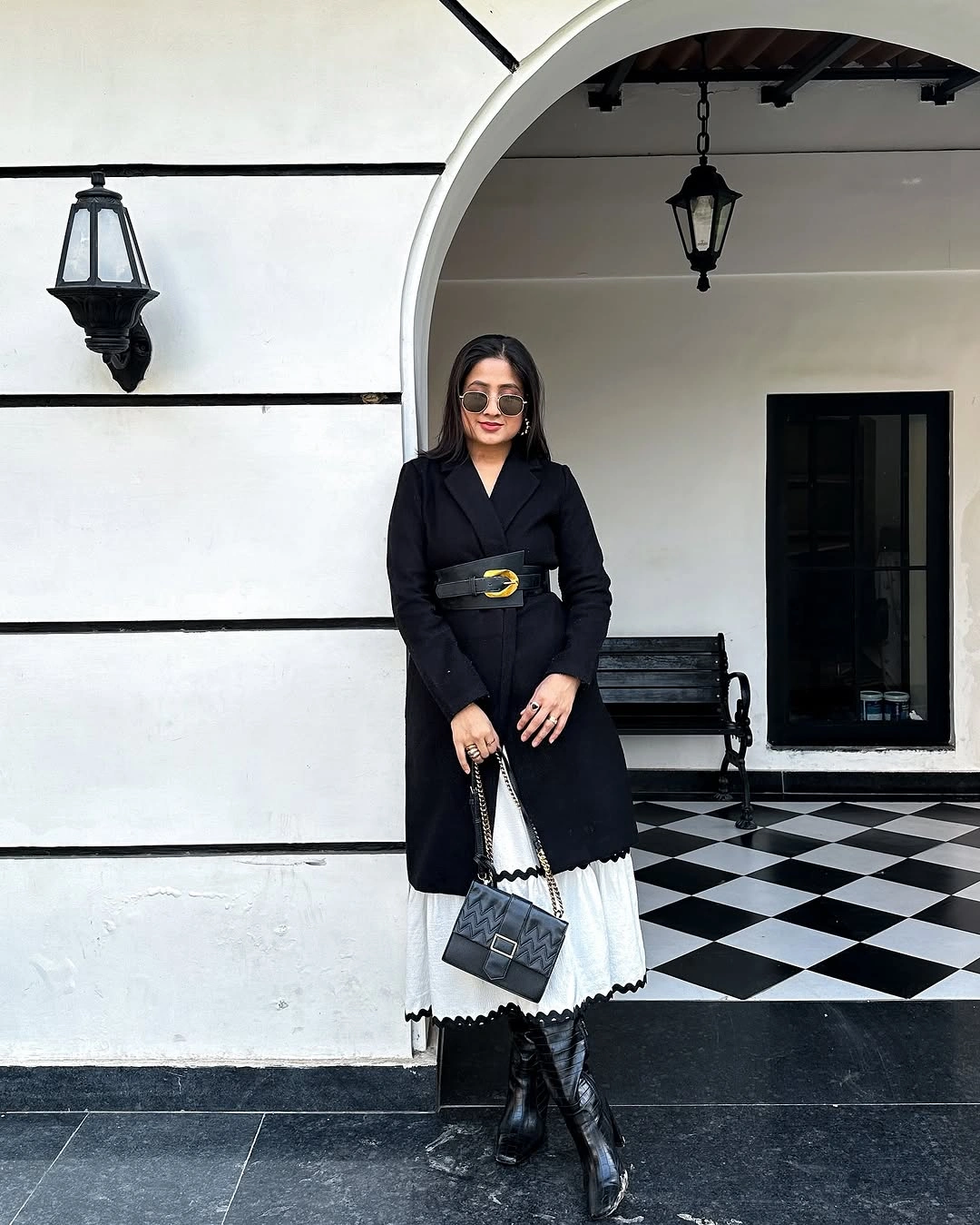 Belted black coat with white scalloped hem, dark boots, and quilted handbag