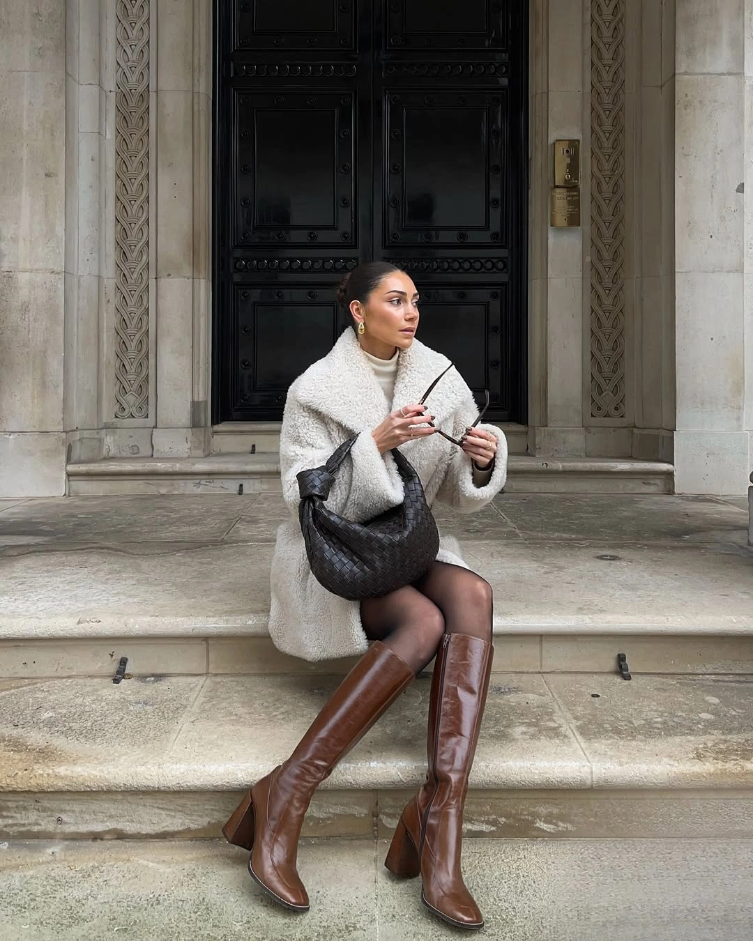 Short cream textured jacket with dark hosiery, tall brown boots, and black bag