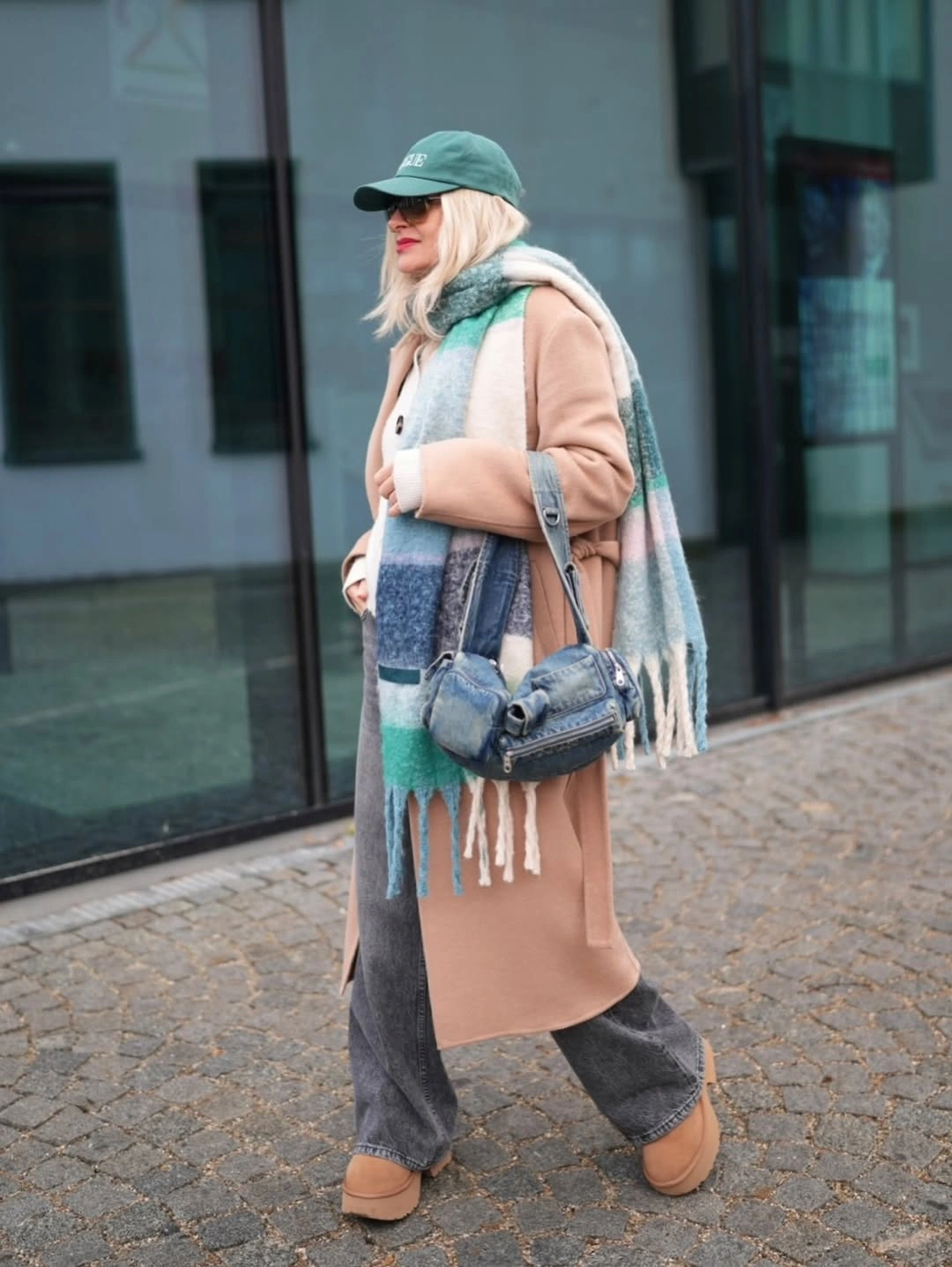 Camel coat with striped blue-green scarf, green cap, gray jeans, and blue bag