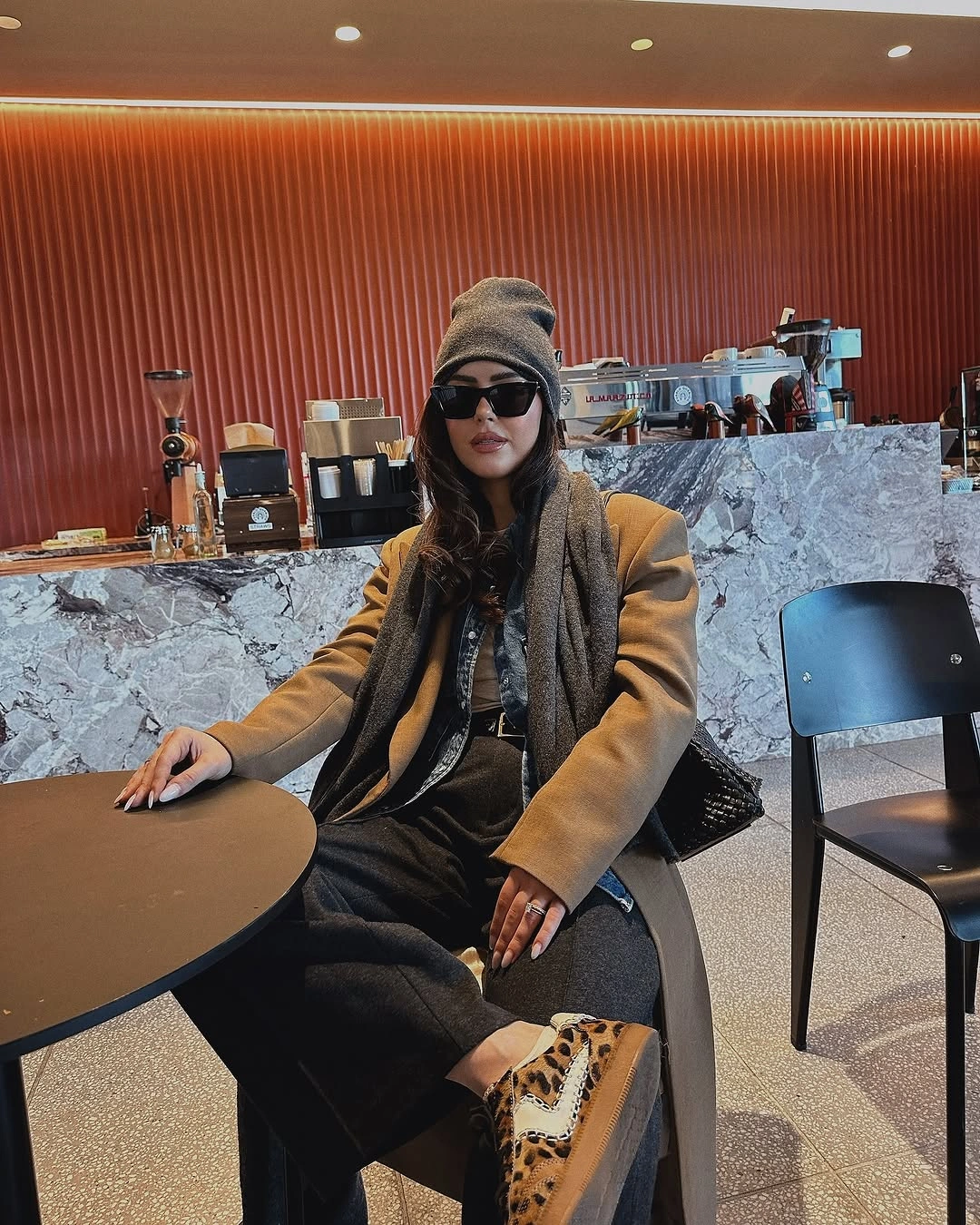 Camel coat and gray scarf with dark trousers and leopard-pattern footwear.