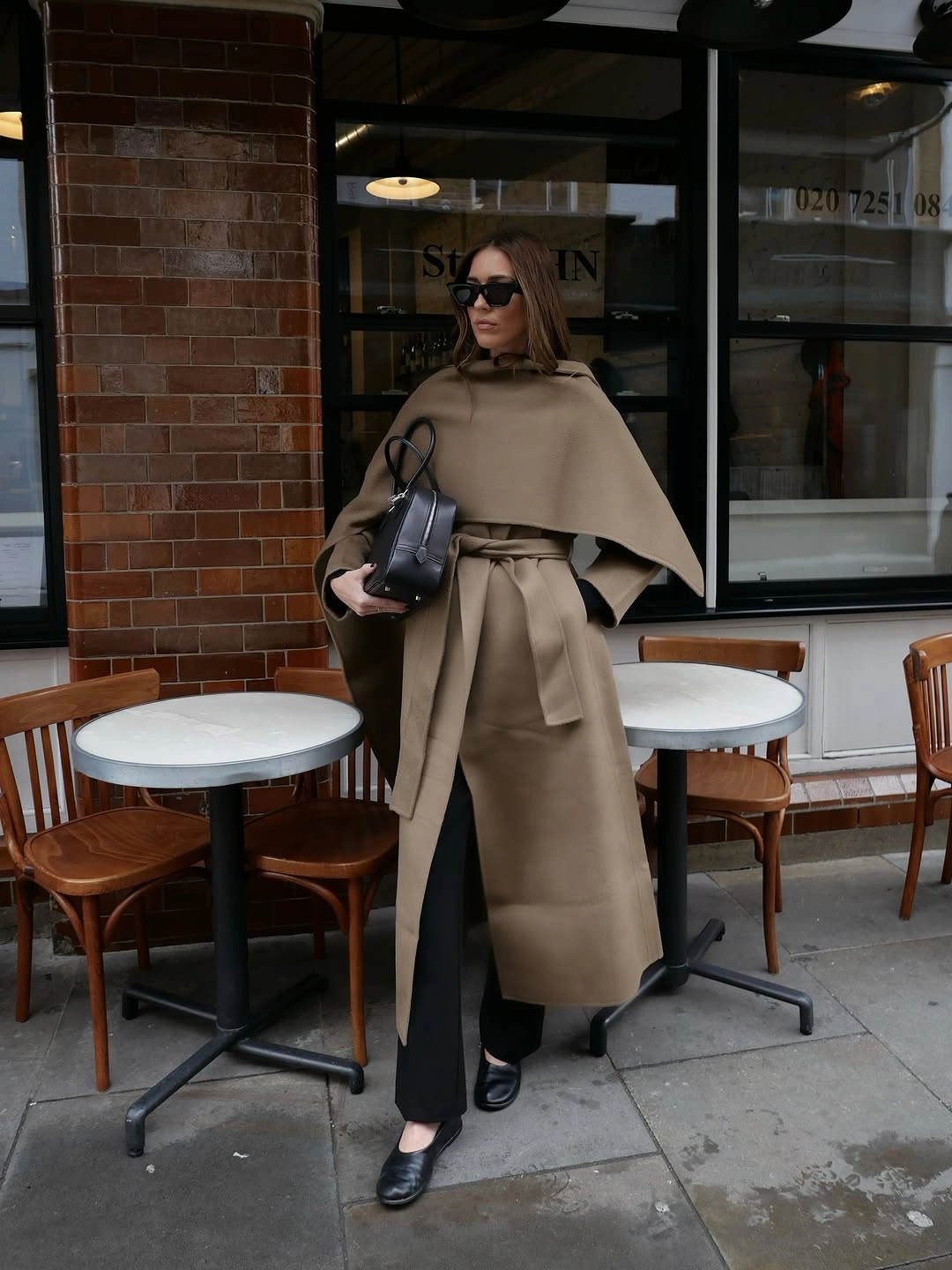 Taupe belted cape-style coat with black trousers, sunglasses, and handbag.