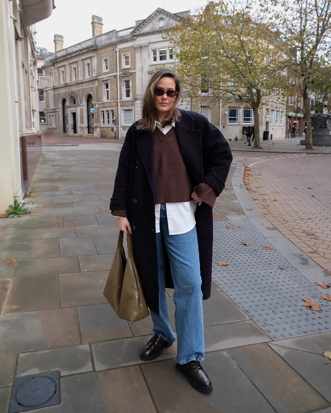Long dark coat over brown knit, white shirt, blue jeans, and dark shoes.