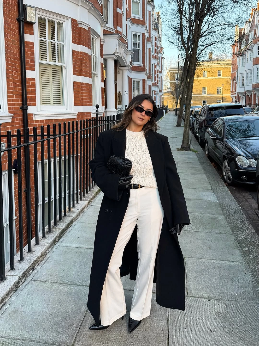 Long black coat over cream sweater, white trousers, and pointed black shoes.