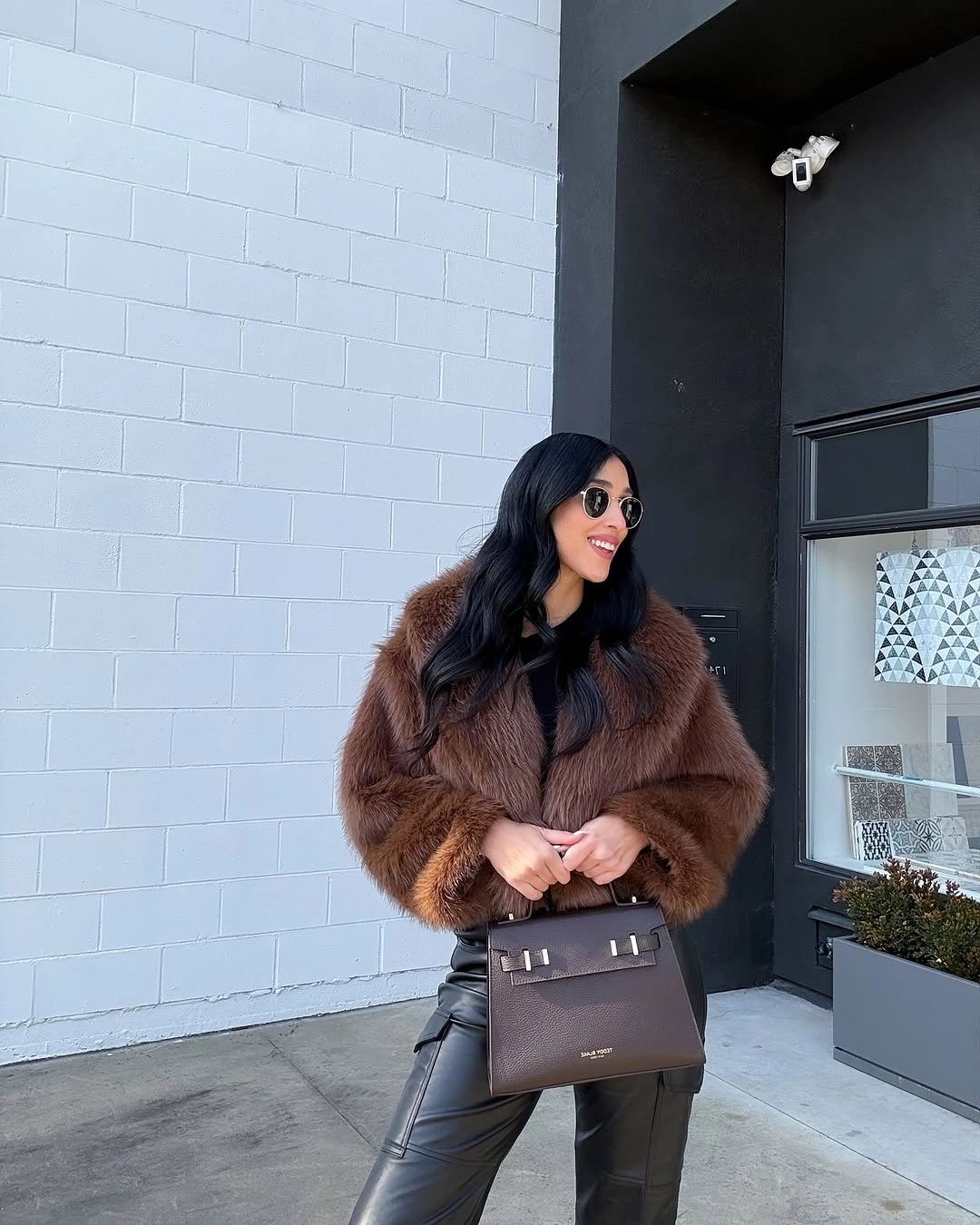 Full chocolate brown plush jacket with glossy black pants and structured handbag